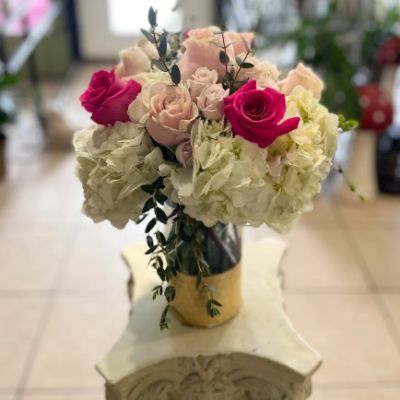 In a keepsake gold bottom vase using hydrangea blooms and shades of pink full size roses and spray roses.