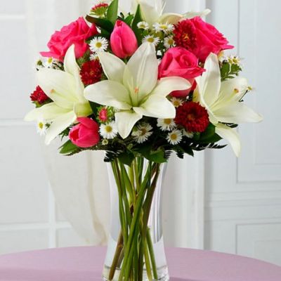 Brilliant fuchsia roses, hot pink tulips, hot pink matsumoto asters, white Asiatic Lilies, white monte casino asters, and lush greens are seated in a clear glass vase.